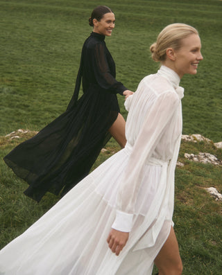 Two women walking outdoors on a grassy field, wearing long sheer high-neck dresses in black and white. The woman in the foreground wears a white dress and is in closer profile, while the woman behind wears a black dress and is further back and more turned toward the camera.