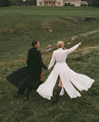 Two women holding hands run across a grassy field toward a large stone mansion in the distance. One wears a flowing black dress and the other wears a long white dress with brown knee-high boots.