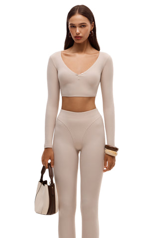 Woman wearing a beige long-sleeve top and leggings holding a handbag on a white background