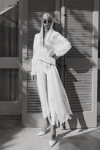 A woman wearing a flowing white long-sleeve wrap-style dress or robe with wide draped sleeves and a loose, airy silhouette, standing in front of shuttered doors in a black-and-white outdoor setting.