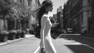 A woman in a fitted beige catsuit walks along a city street, holding a dark textured handbag. The photo is in black and white with a blurred urban background.